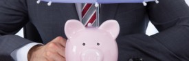 Businessman Sheltering Piggybank With Umbrella At Desk