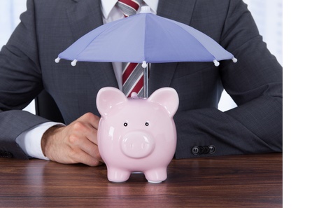 Businessman Sheltering Piggybank With Umbrella At Desk